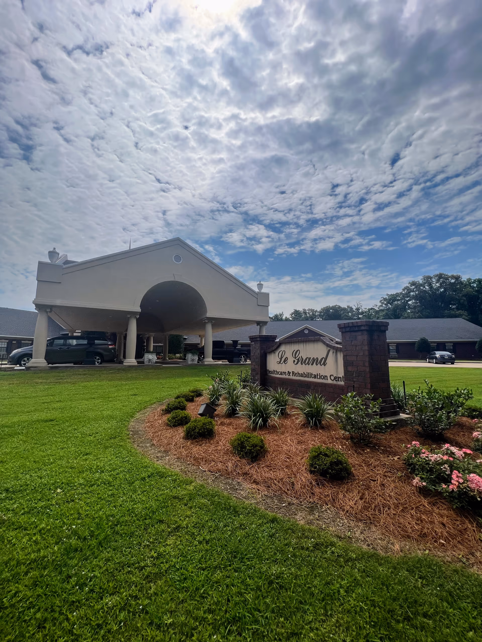 Front entrance and porte-cochère of Le Grand Healthcare & Rehabilitation Center with a landscaped sign under a cloudy sky.