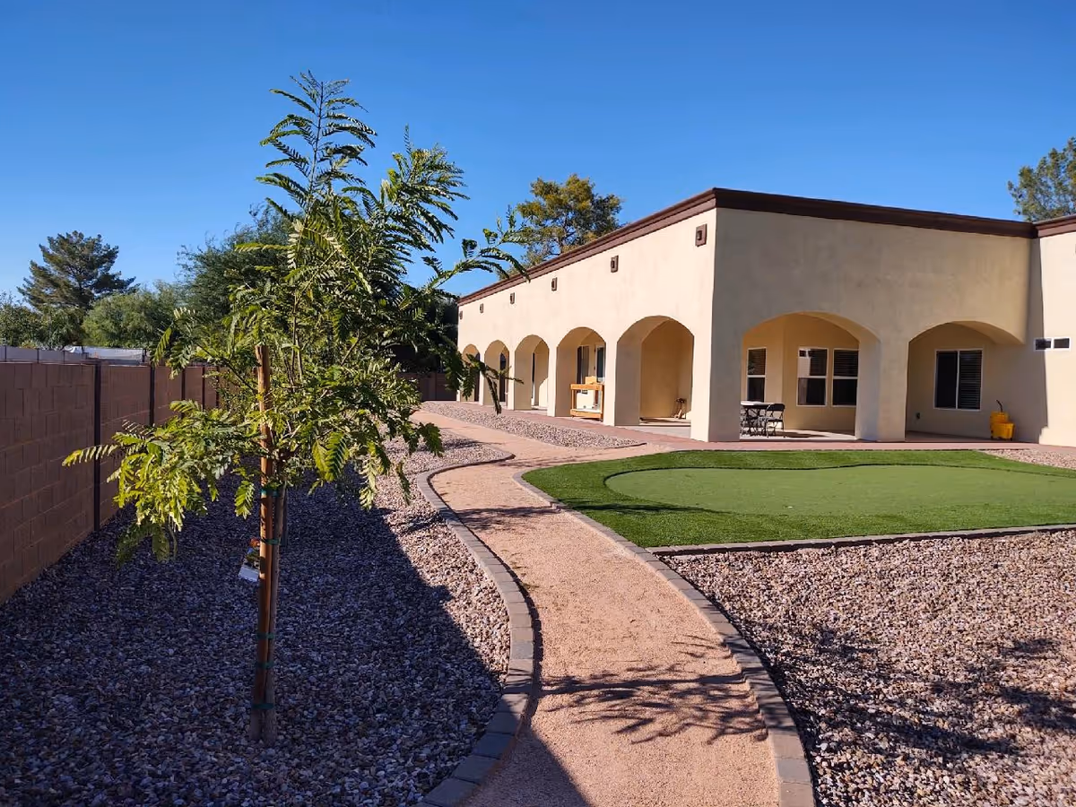 Outdoor view of a senior living facility with a curved gravel pathway bordered by small trees and rocks, leading to a beige building with arched doorways and windows under a clear blue sky.