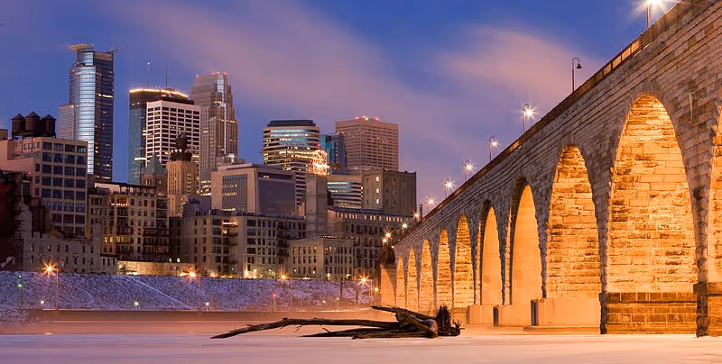 Dusk cityscape with a stone-arched bridge and illuminated skyscrapers.