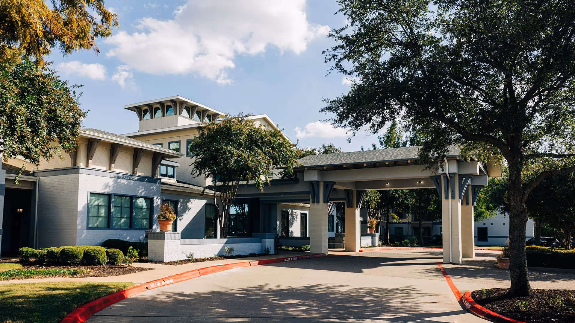 Exterior view of Parkview in Allen senior living facility showing a covered entrance with columns, surrounded by trees and landscaping under a partly cloudy sky.