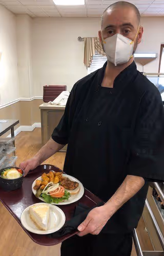 A man wearing a black chef uniform and a white face mask is holding a tray with a plate of food including fried items, a salad, and a slice of pie in a dining or kitchen area with beige walls and a window with blinds and a curtain.