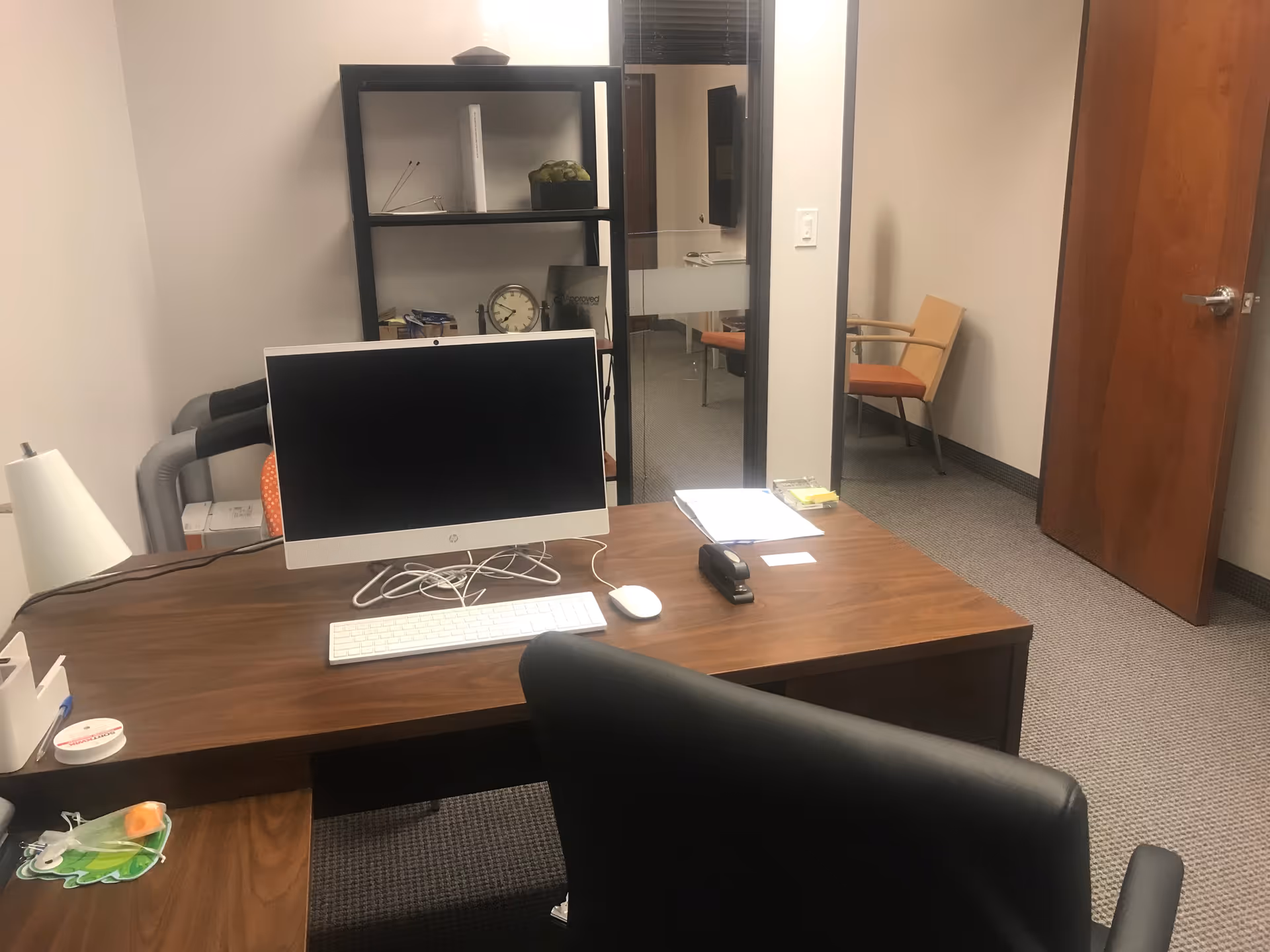Office workspace with computer, desk, shelves, and a seating area visible through doorway.