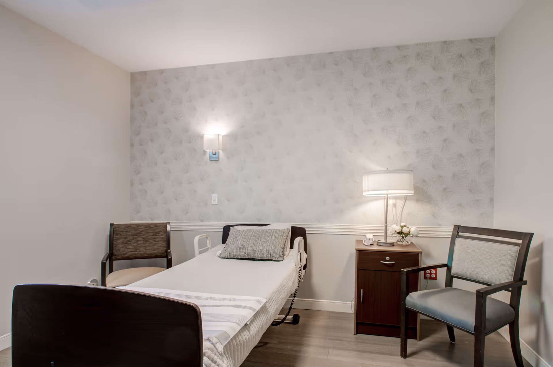 A single hospital-style bedroom features a bed, two chairs, and a bedside cabinet.