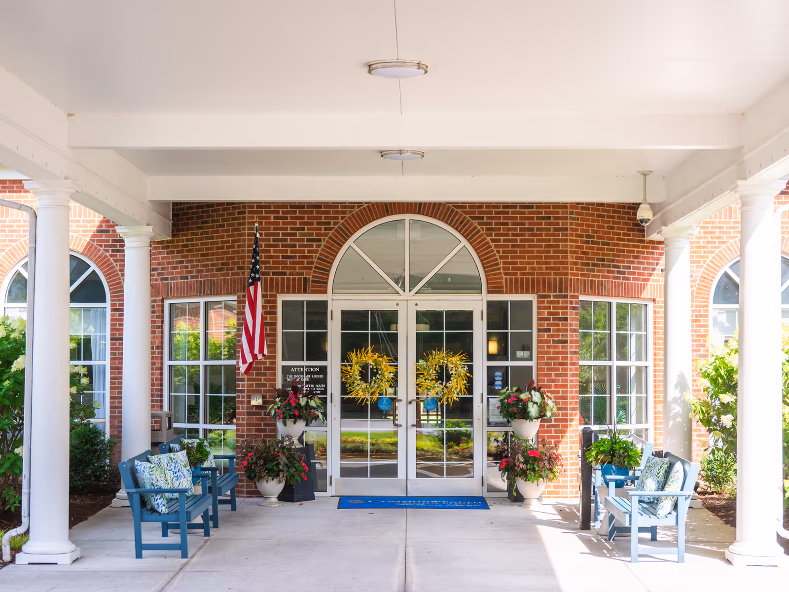 Brick building entrance with white columns, glass doors, wreaths, seating, plants, and an American flag.