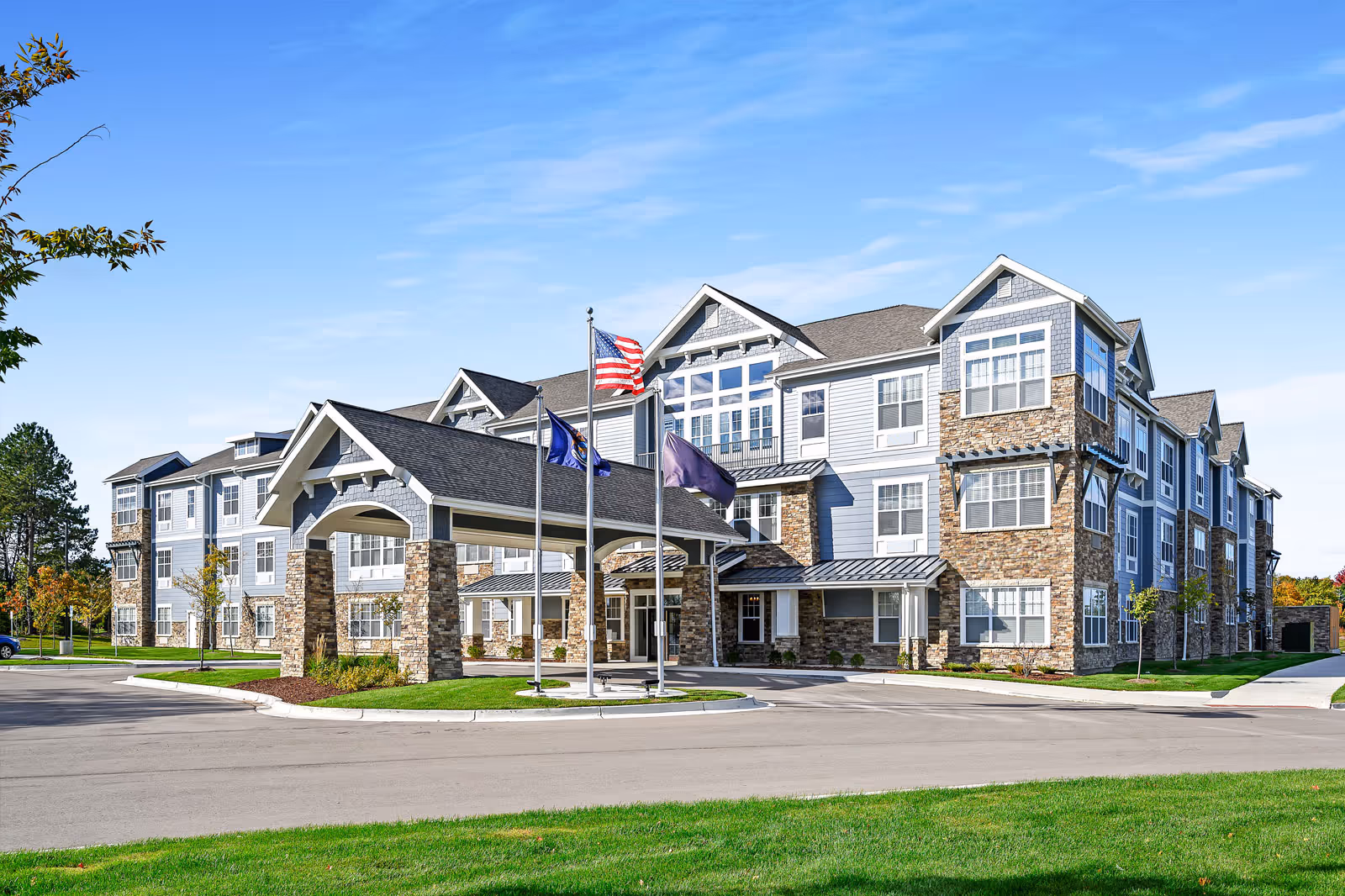 Exterior view of a large, modern senior living facility building with stone and siding facade, multiple windows, and a covered entrance with three flagpoles displaying flags. The building is surrounded by a paved driveway and green lawn under a clear blue sky.
