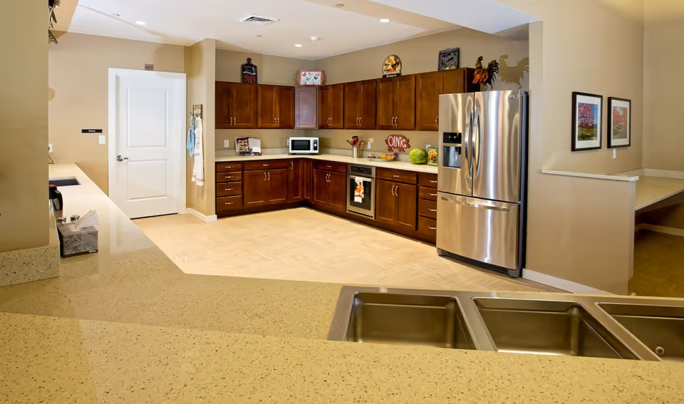 Spacious communal kitchen with stainless steel refrigerator, wooden cabinets, microwave, and a large countertop with sinks in the foreground.