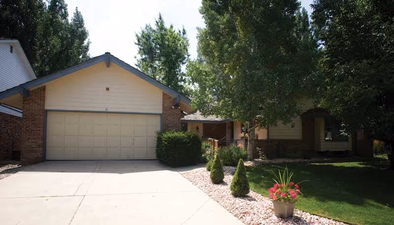 Exterior view of a single-story assisted living facility building with a two-car garage, surrounded by trees and landscaping including small bushes and a flower pot with pink flowers near the driveway.