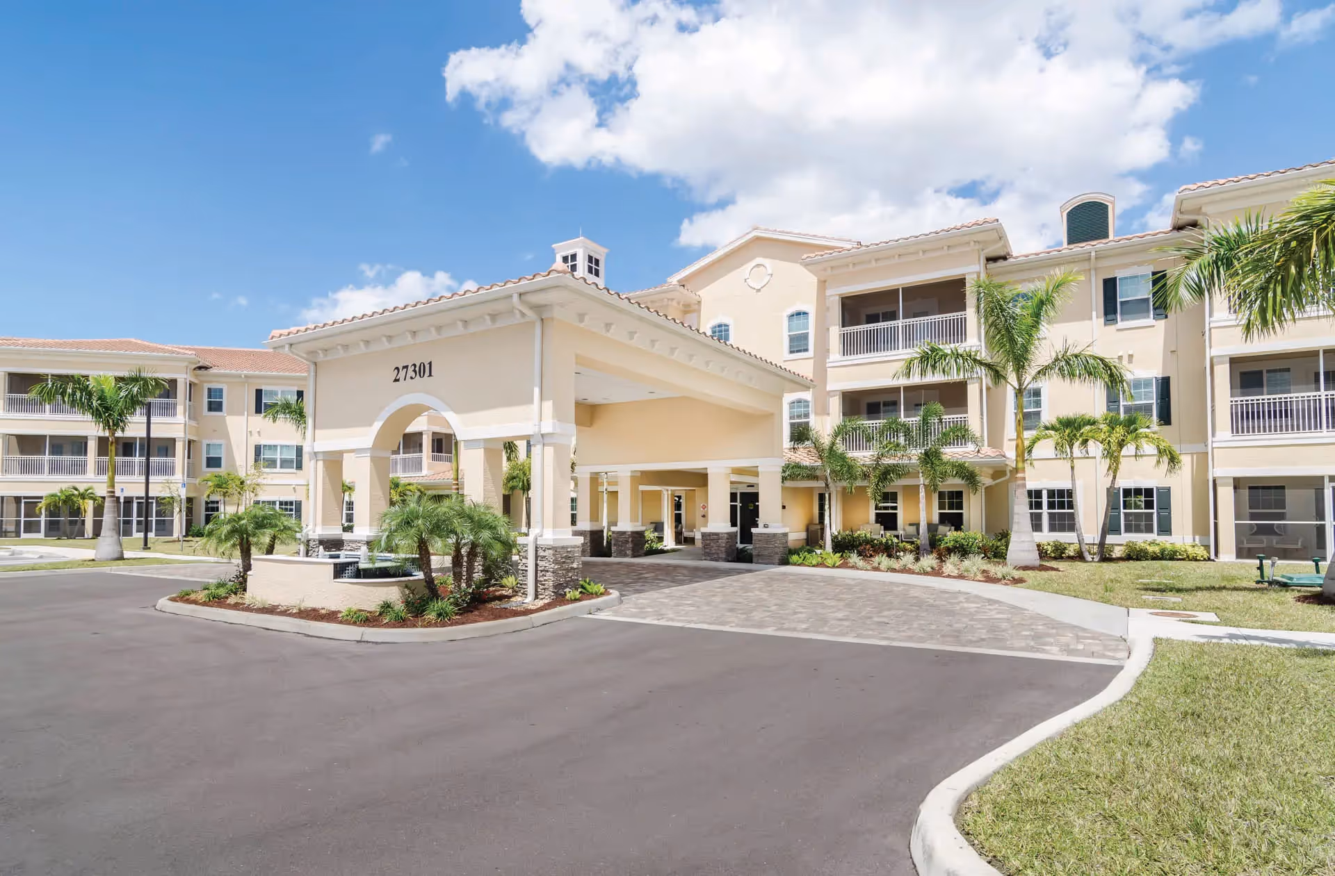 Front entrance with a porte-cochère of a three-story beige senior living building surrounded by palm trees and landscaping, showing the address number 27301.