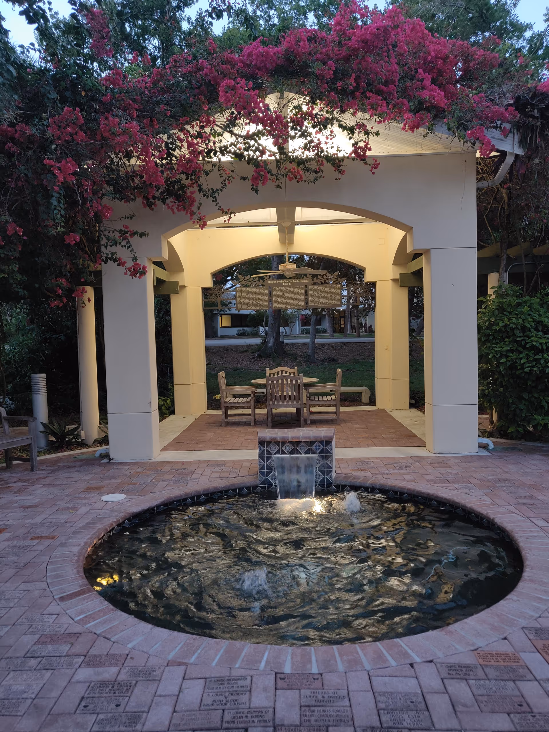 Outdoor courtyard with a circular fountain, brick paving, and seating beneath flowering arches.