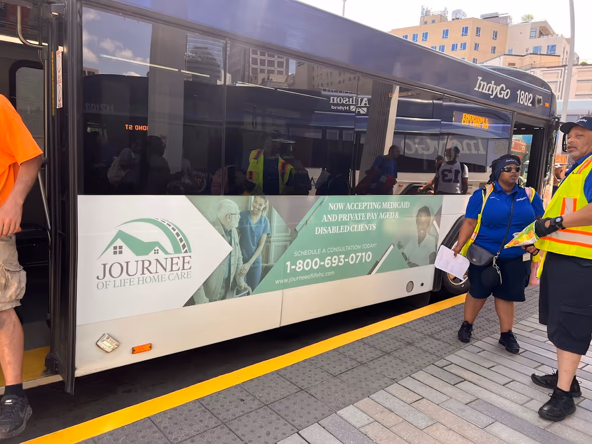 A bus with Journee of Life Home Care advertisement beside workers at a curb.