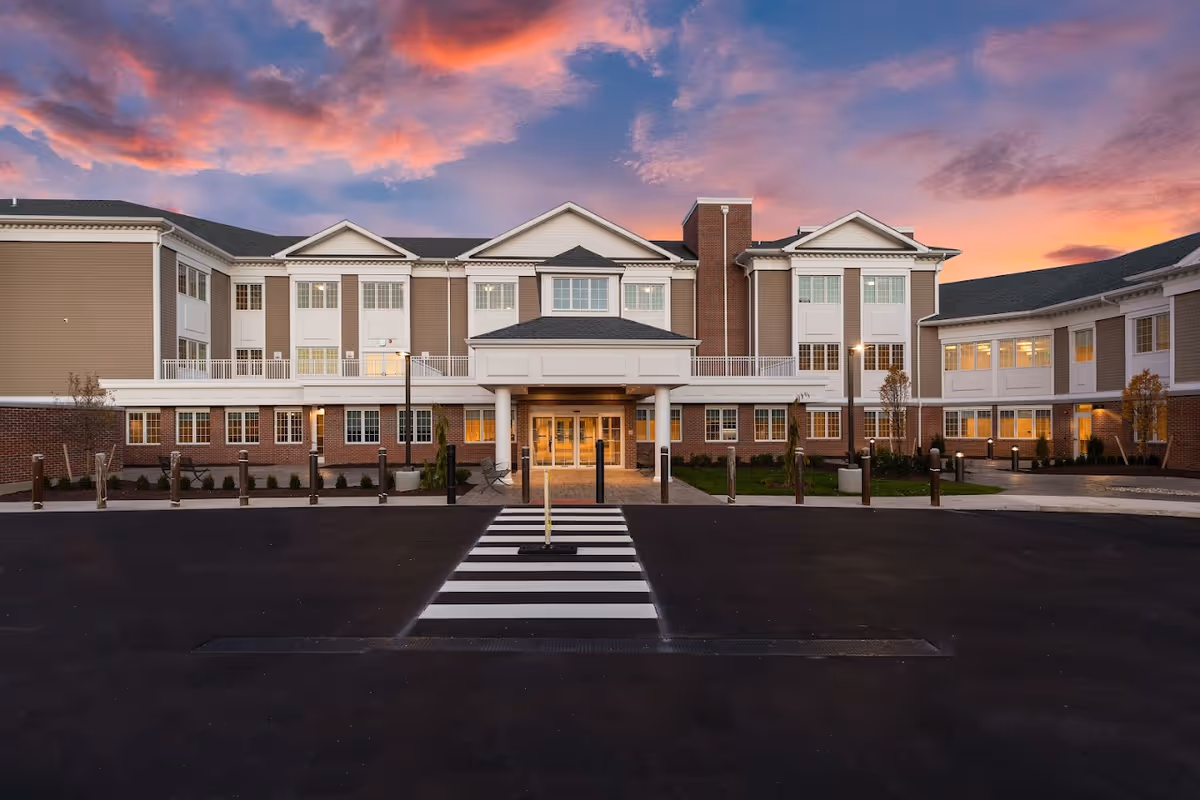Front exterior view of a large senior living facility building with multiple windows, a covered entrance with columns, and a crosswalk leading to the entrance. The sky is colorful with a sunset featuring pink and purple clouds.