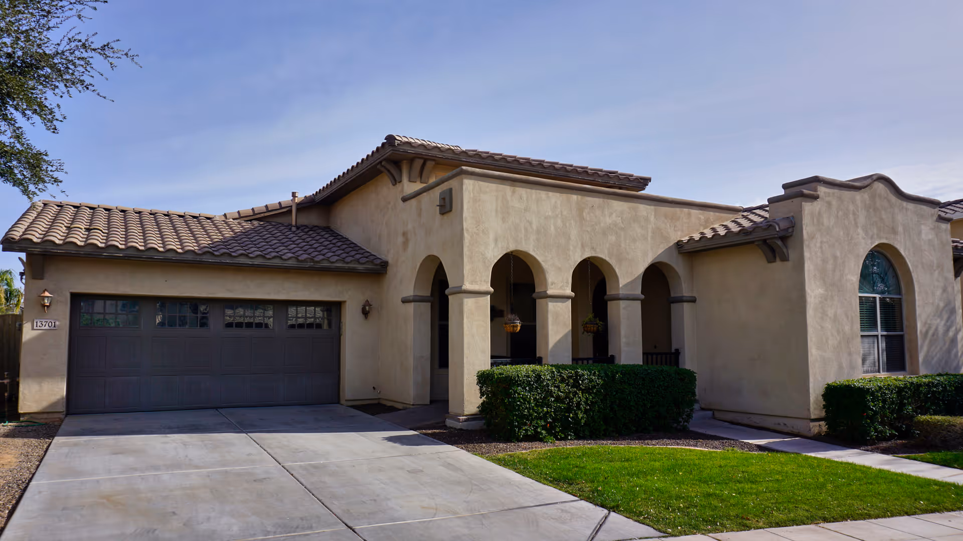 Exterior view of a single-story beige stucco house with a tiled roof, a two-car garage, arched front porch supported by columns, and neatly trimmed bushes and green lawn in front.