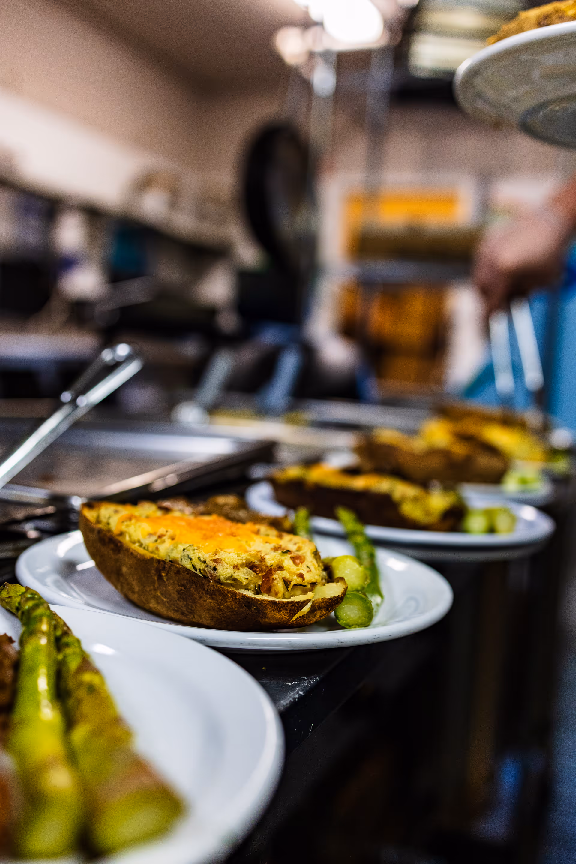 Close-up view of plates with stuffed baked potatoes and asparagus spears lined up on a kitchen counter, with a person in the background serving food.
