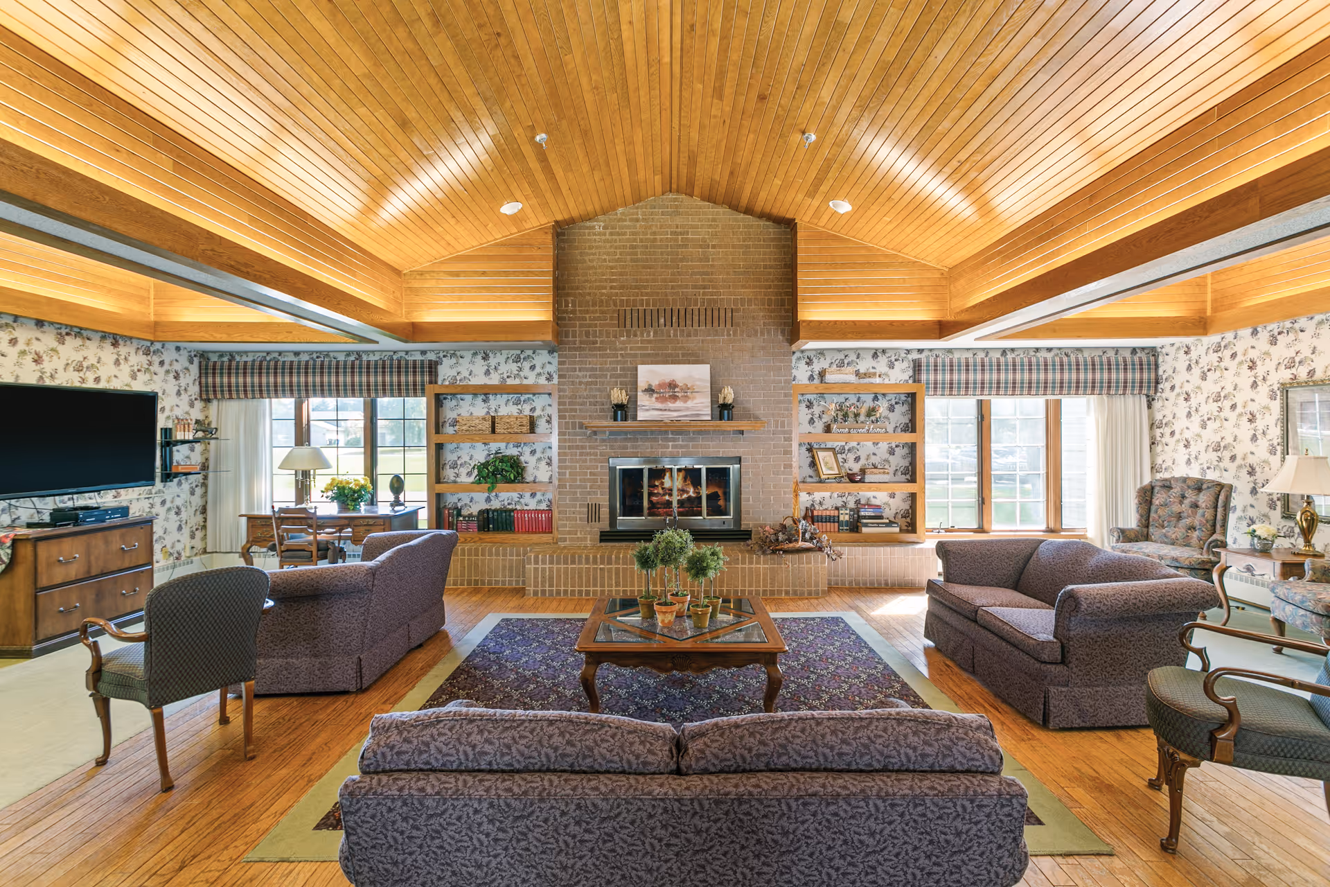 A spacious living room with a high wooden ceiling and a large brick fireplace in the center. The room features floral wallpaper, multiple windows with plaid valances, and a variety of seating including sofas, armchairs, and a coffee table with plants. There is a large flat-screen TV on the left side and bookshelves flanking the fireplace.