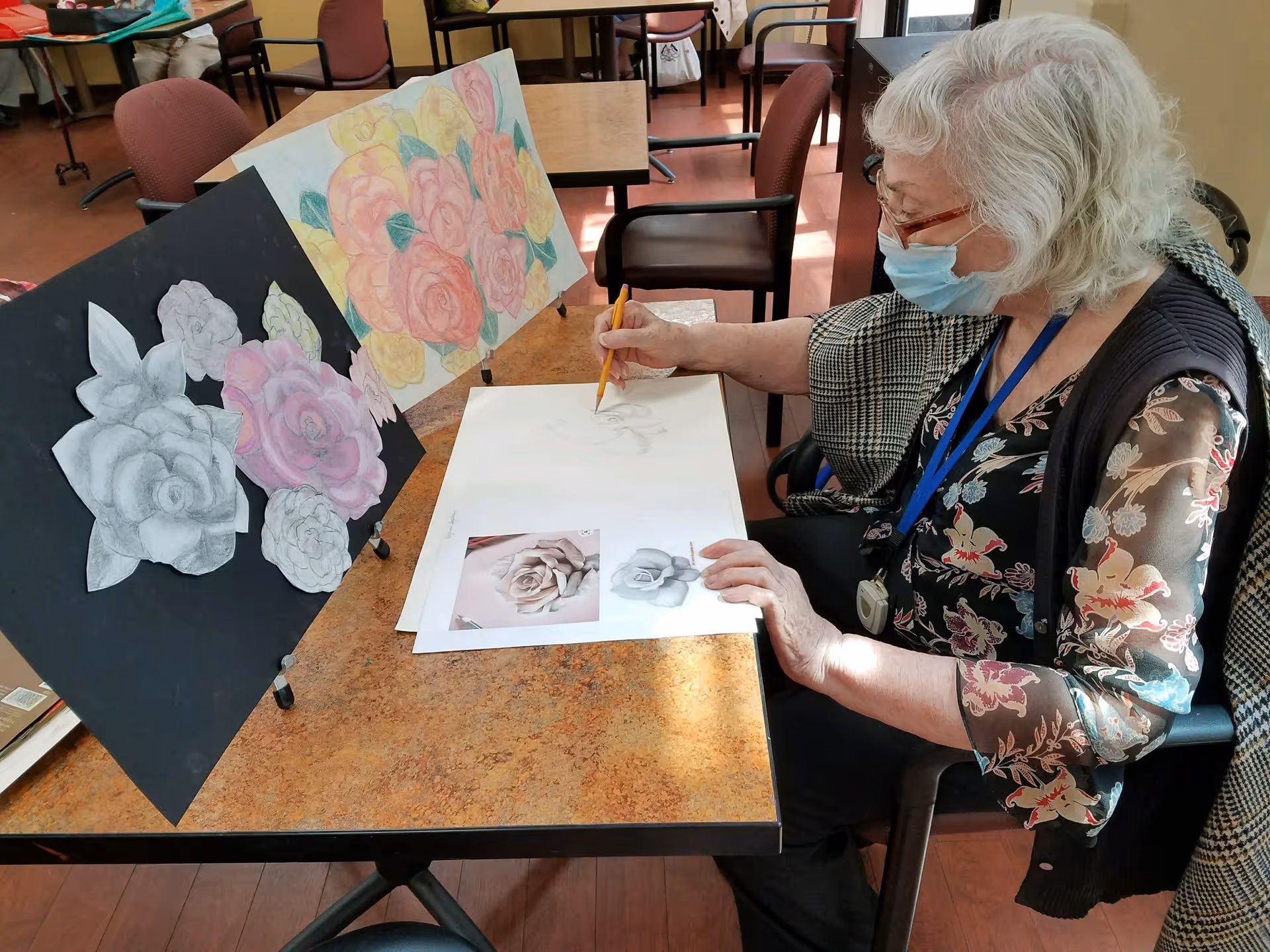 An elderly woman wearing a face mask and glasses is seated at a table, drawing a rose on a large sheet of paper. She is using a pencil and referencing two printed images of roses. On the table, there are two boards displaying colored and black-and-white drawings of roses. The setting appears to be a communal indoor space with chairs and tables.