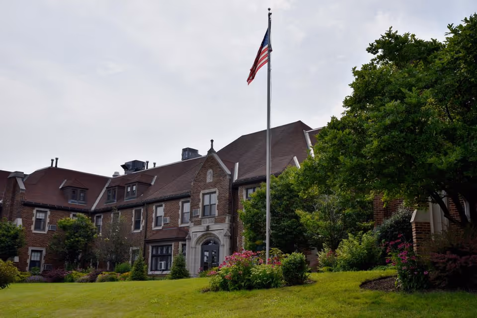 Brick senior living building with flagpole, manicured lawn, and trees in front.