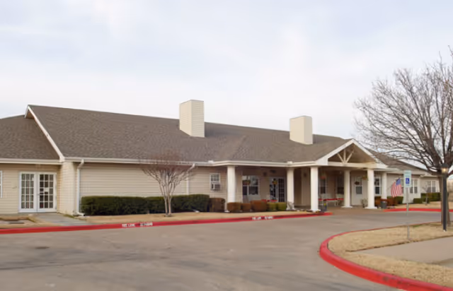 Single-story senior living building with a covered entrance, circular driveway, and sparse landscaping under a cloudy sky.