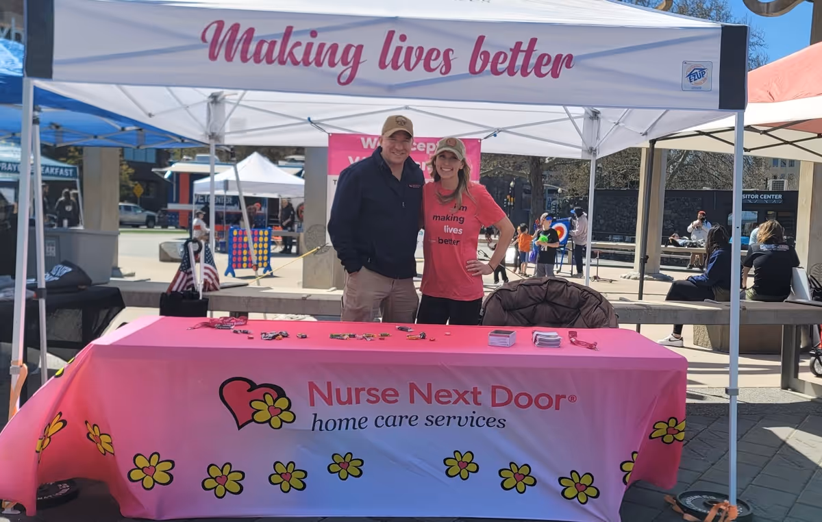 Two people stand beside a pink Nurse Next Door table at an outdoor event.