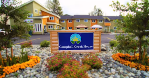 View of the front exterior of Campbell Creek House, featuring a landscaped garden with colorful flowers and rocks surrounding a wooden sign with the facility's name. The building is a multi-story structure painted in green and orange tones.
