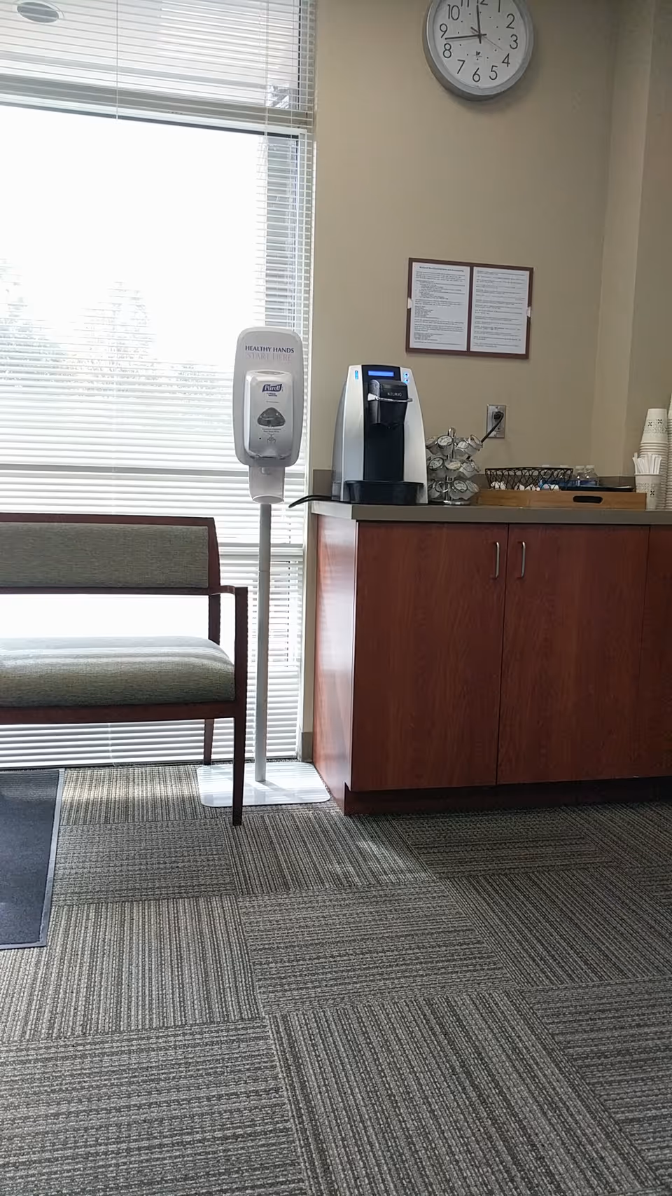 Interior view of a clinic reception area with a bench, sanitizer stand, coffee machine, clock.