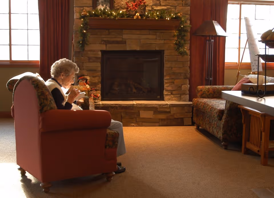 An elderly woman sitting in a cushioned armchair in a cozy living room with a stone fireplace decorated with garland and holiday ornaments. The room has large windows with red curtains, a patterned sofa, a floor lamp, and a table with decorative items.