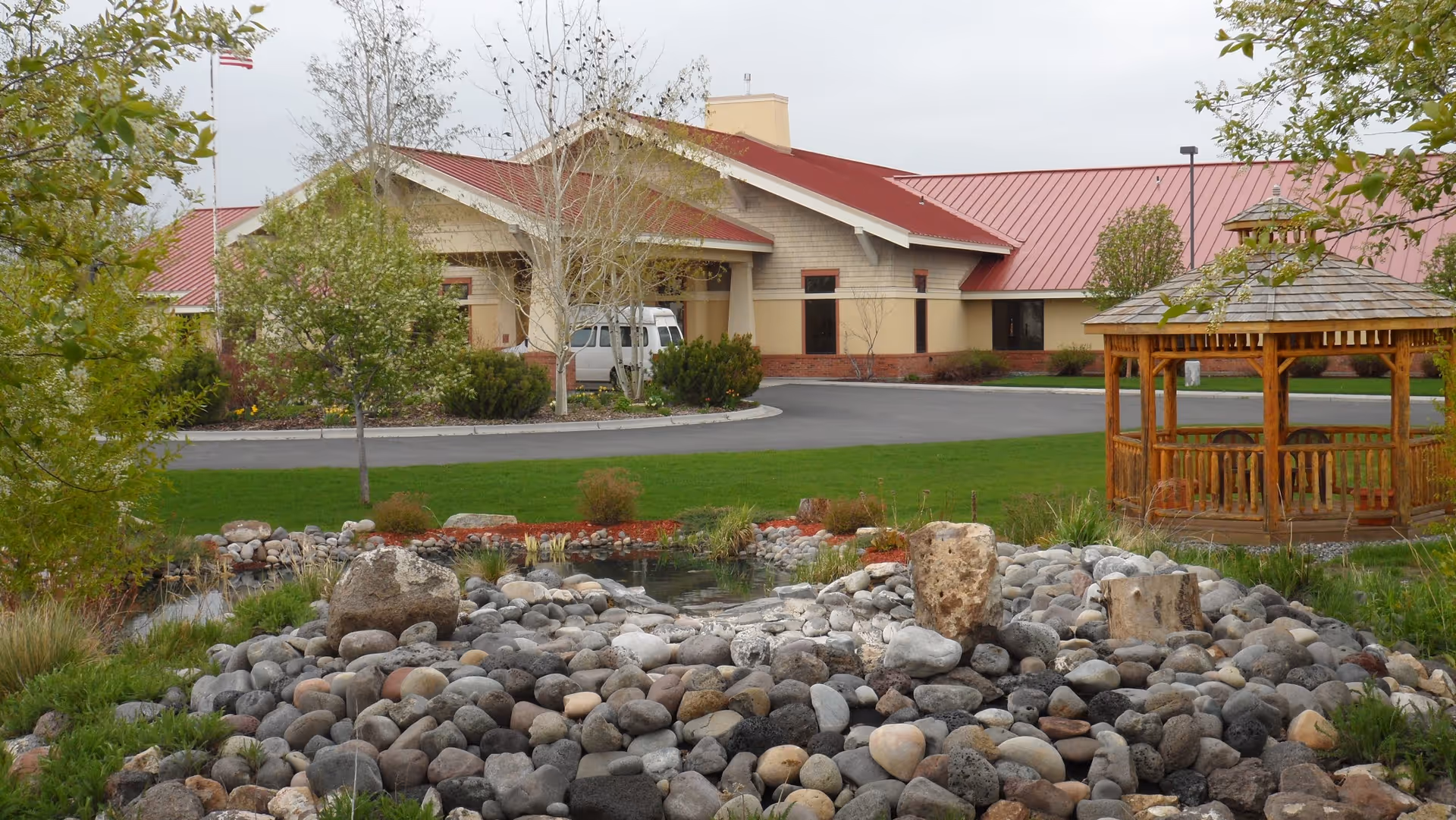 Front exterior of a single-story building with a red metal roof, driveway entrance, wooden gazebo, and a landscaped rock pond in the foreground.