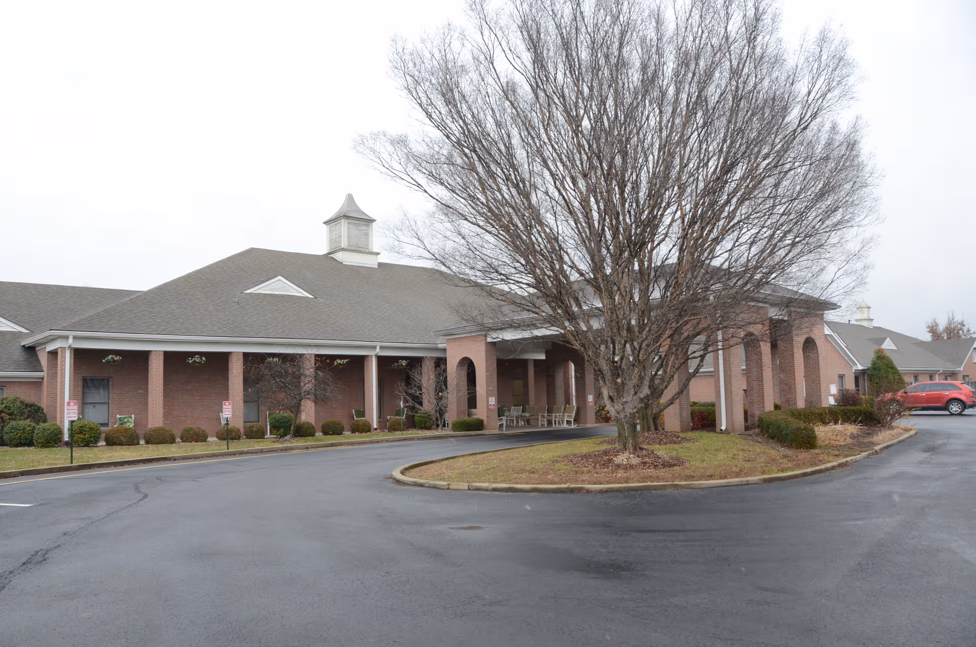 Brick senior living facility with circular driveway and leafless tree at entrance.