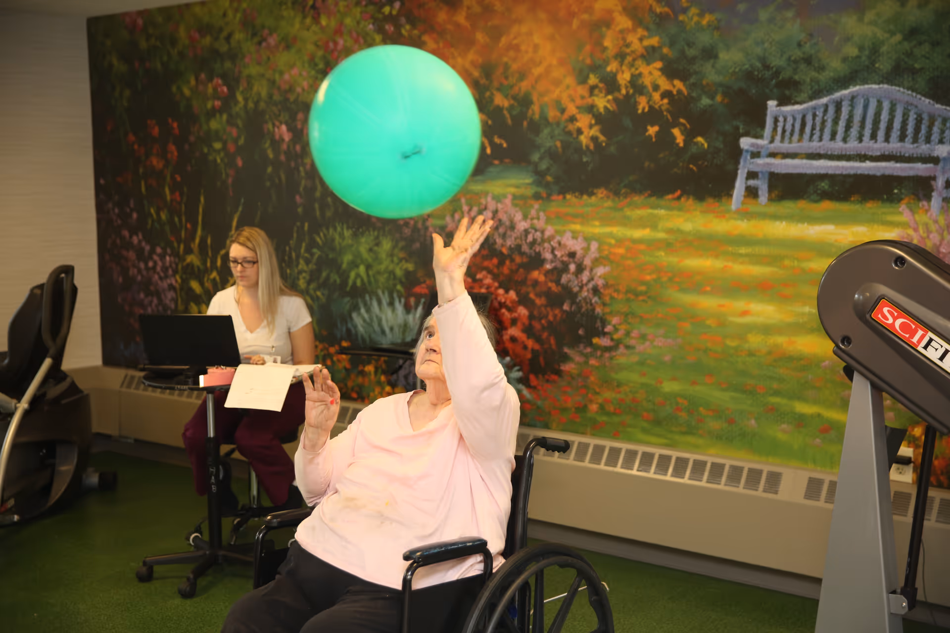 An elderly woman in a wheelchair playing with a large green exercise ball indoors. A young woman is seated nearby working on a laptop with papers on a small table. The background features a colorful mural of a garden scene with flowers and a bench.