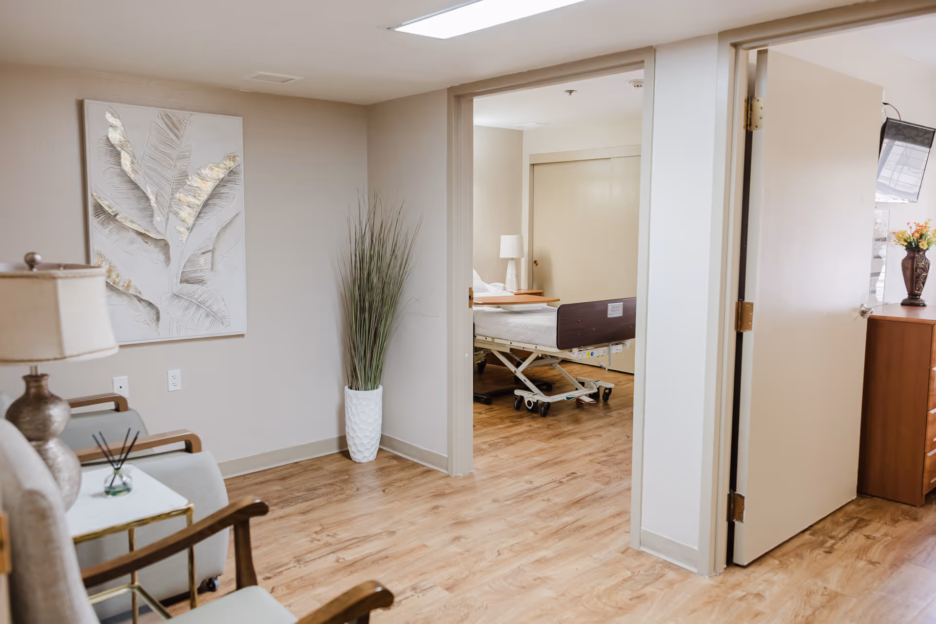 Living room with beige walls, furniture, a tall plant, and a patient room beyond.