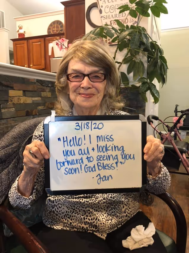 An elderly woman with glasses and gray hair is sitting in a chair indoors, holding a whiteboard with a handwritten message dated 3/18/20 that says, 'Hello!! I miss you all + looking forward to seeing you soon! God Bless! - Jan'. Behind her is a stone wall, a large green plant, and a walker is visible to the right.