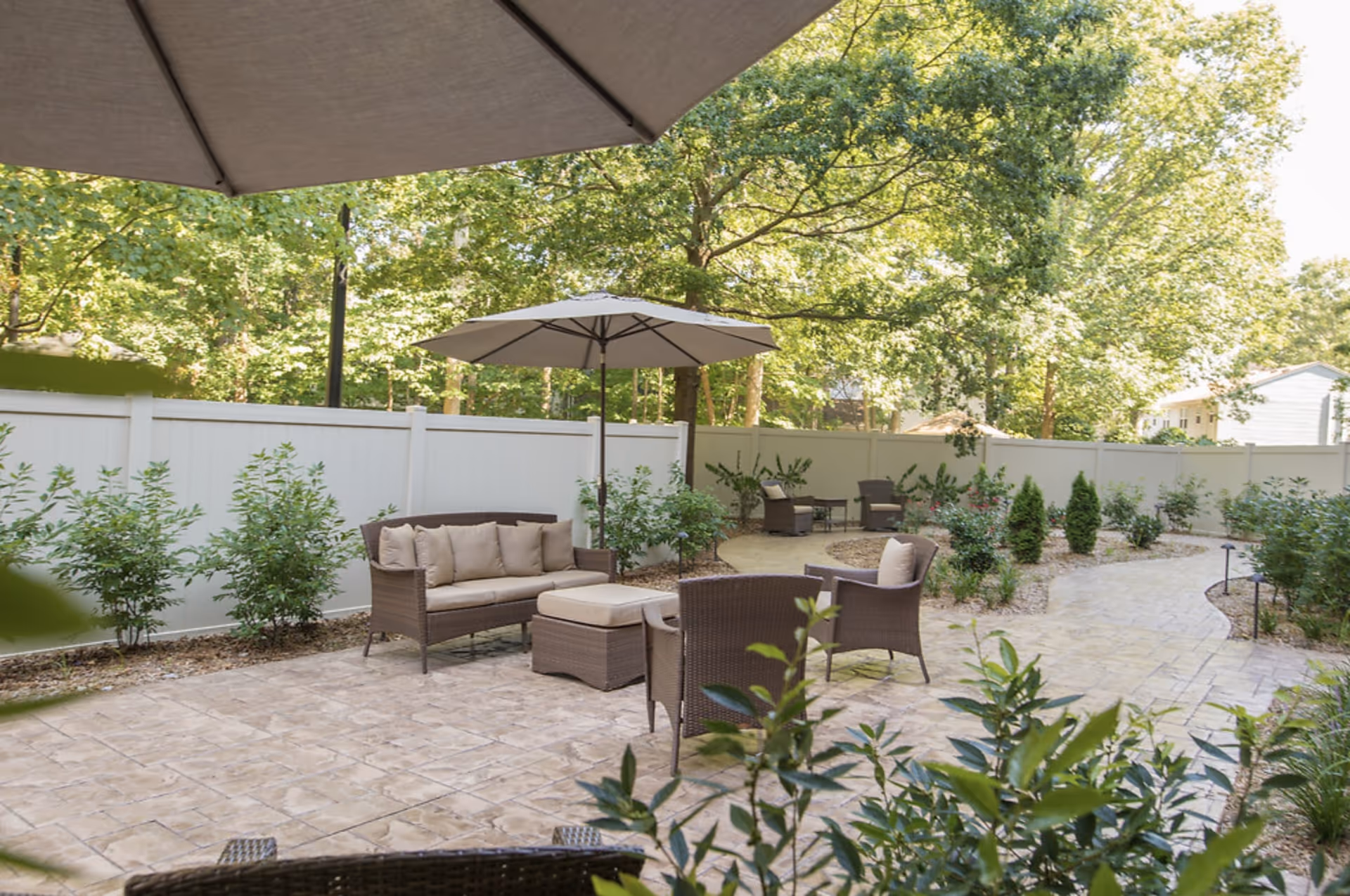 Outdoor patio area with wicker furniture including a sofa, chairs, and an ottoman under large umbrellas, surrounded by greenery and a white fence in the background.