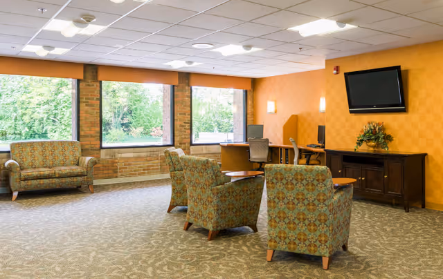 A bright and spacious common area with large windows showing greenery outside. The room features patterned armchairs and a loveseat, a wooden cabinet with a flat-screen TV mounted above it, and a desk area with two computer workstations and chairs. The walls are painted orange with a subtle checkered pattern, and the carpet has a decorative design.