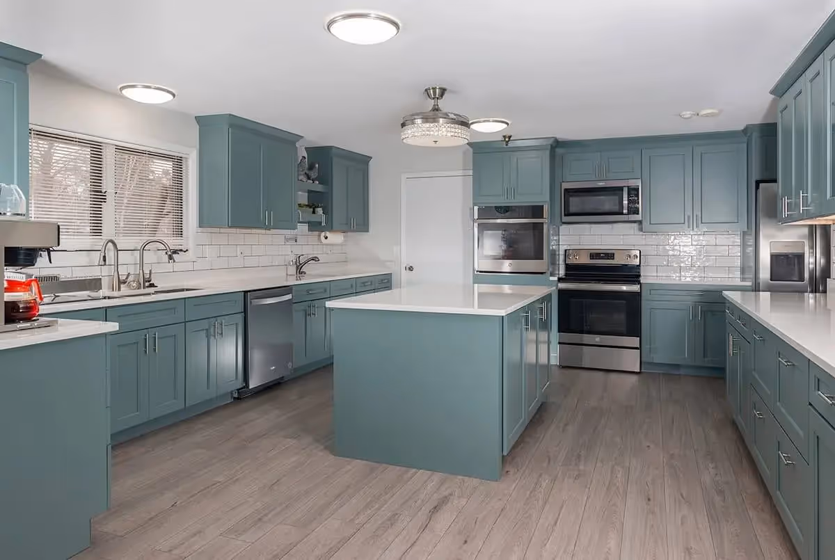A spacious modern kitchen with teal cabinets and drawers, white countertops, and a central island. The kitchen features stainless steel appliances including a refrigerator, oven, microwave, and dishwasher. There is a coffee maker on the counter near a window with blinds. The floor is wood-style laminate, and the ceiling has multiple light fixtures.