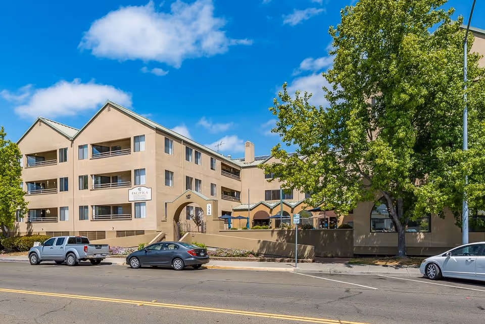 Exterior view of a multi-story senior living facility building with beige walls and green roofs under a blue sky with some clouds. There are several cars parked along the street in front of the building, and a large tree partially obscures part of the building on the right side.