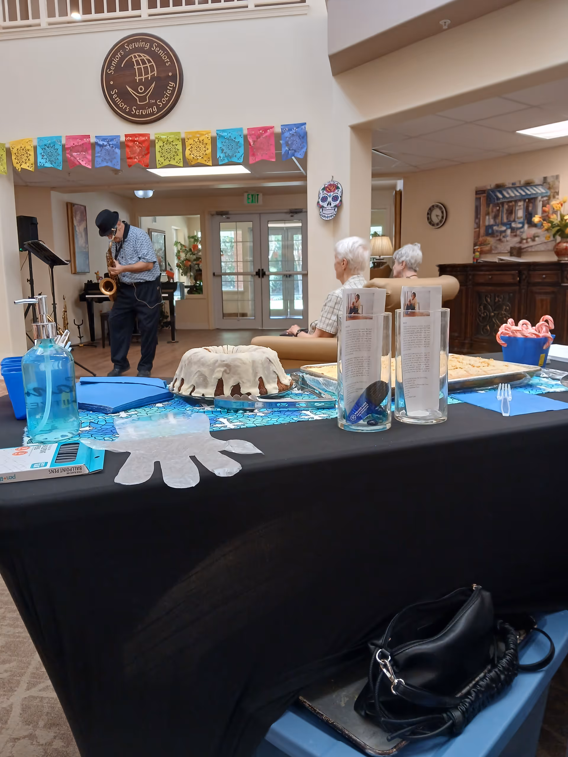 Saxophonist performs at a senior living facility event beside cake table.