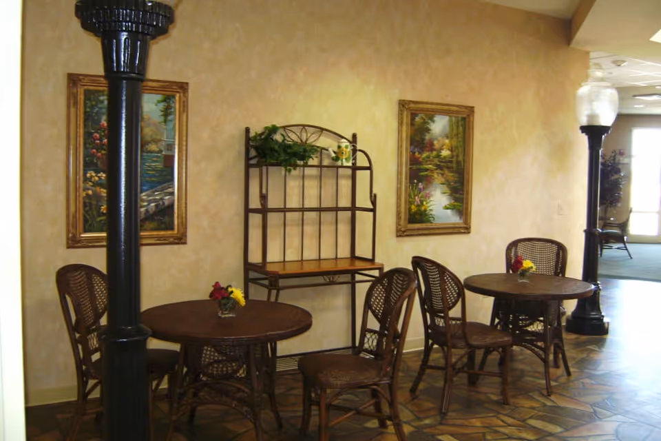 Interior seating area with two round wooden tables, each surrounded by three wicker chairs. The walls are beige with two framed landscape paintings. A metal shelf with a plant and decorative items is mounted on the wall between the paintings. Two black lamp posts with glass light fixtures are positioned near the tables. The floor has a patterned tile design, and there is a doorway leading to another room in the background.