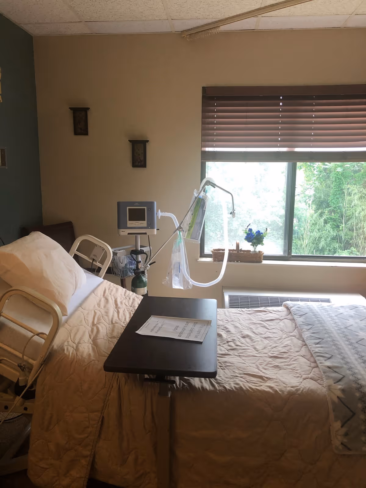 A hospital-style bed with beige bedding and a pillow in a room with a window covered by brown blinds. There is a medical monitor and oxygen tank next to the bed, and an overbed table with a paper on it. A small basket with blue flowers is on the windowsill.