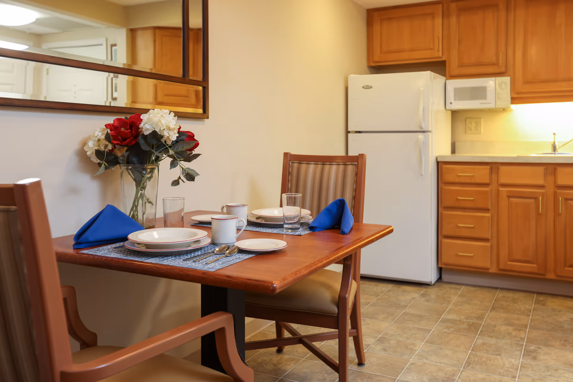 A small dining area with a wooden table set for two, including plates, cups, glasses, and blue folded napkins. There is a vase with red and white flowers on the table. In the background, a kitchen area with wooden cabinets, a white refrigerator, a microwave, and a countertop with a sink is visible. A large mirror hangs on the wall above the table.