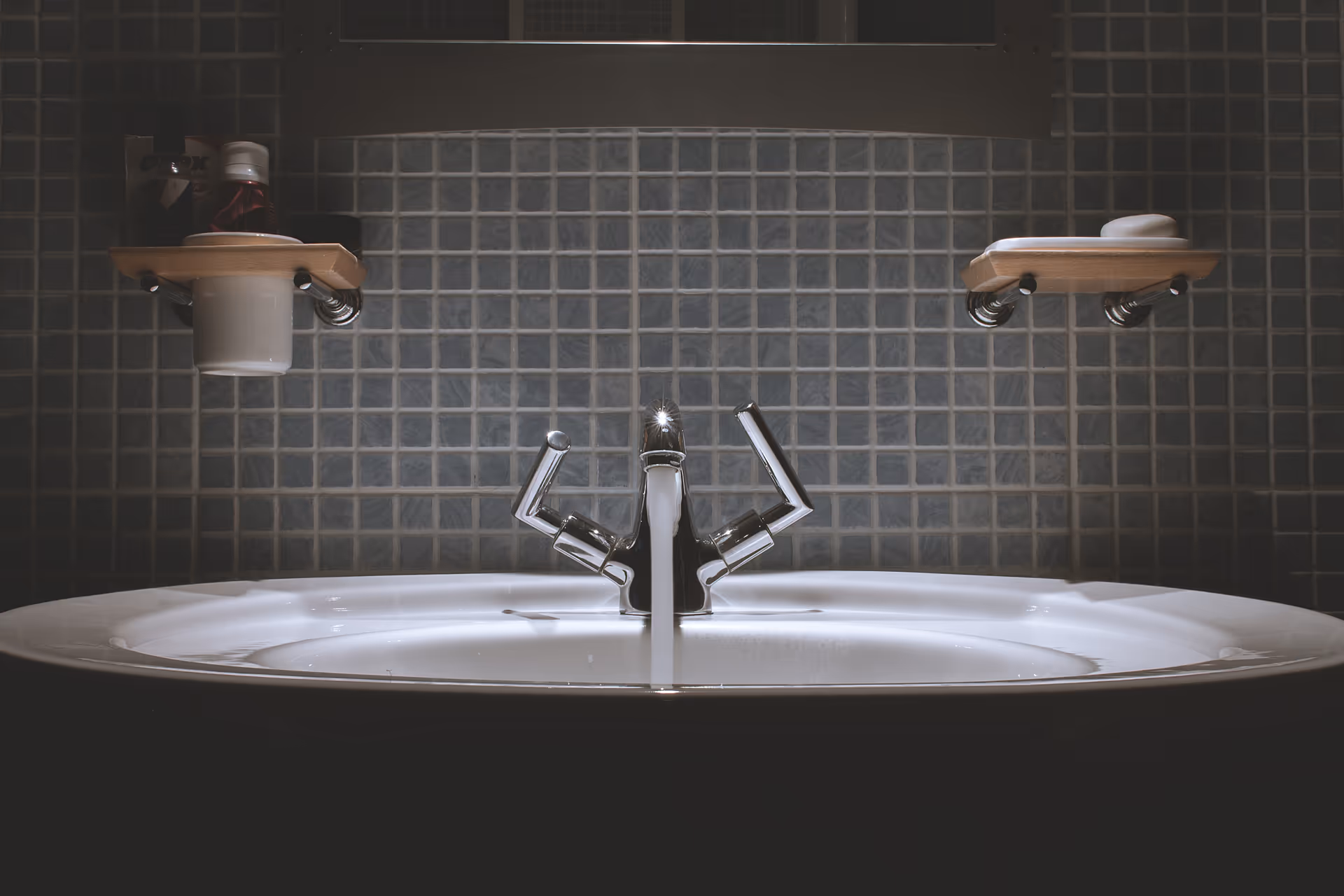 Bathroom sink with dual faucets, tiled wall, and shelves.