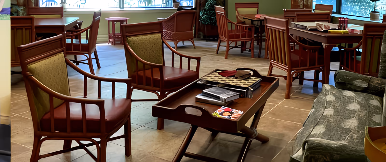 Cozy communal lounge with wooden chairs, tables, and a coffee table holding books and magazines near large windows.