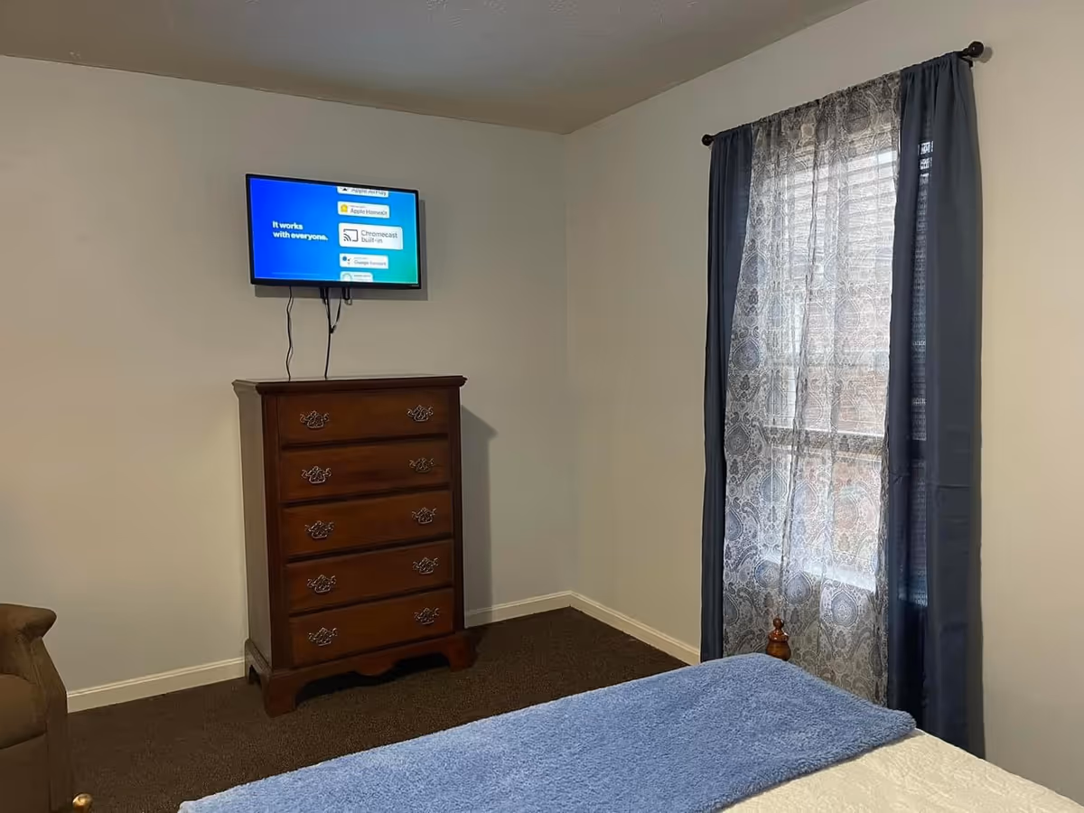 A bedroom with a wooden dresser, wall-mounted TV, window with lace curtains, and a bed.