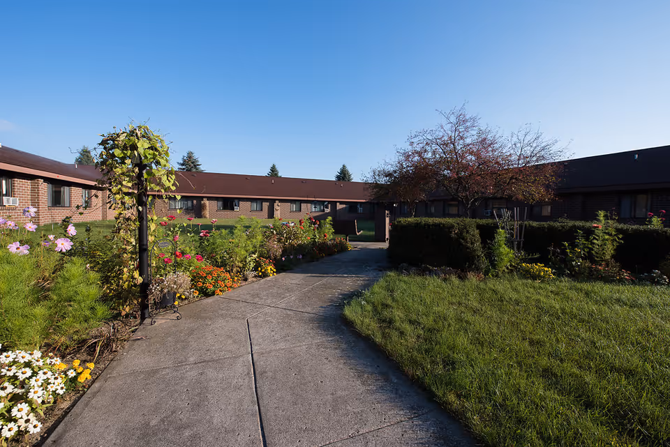Concrete pathway through a landscaped courtyard with flower beds leading to a single-story brick apartment building under a clear blue sky.