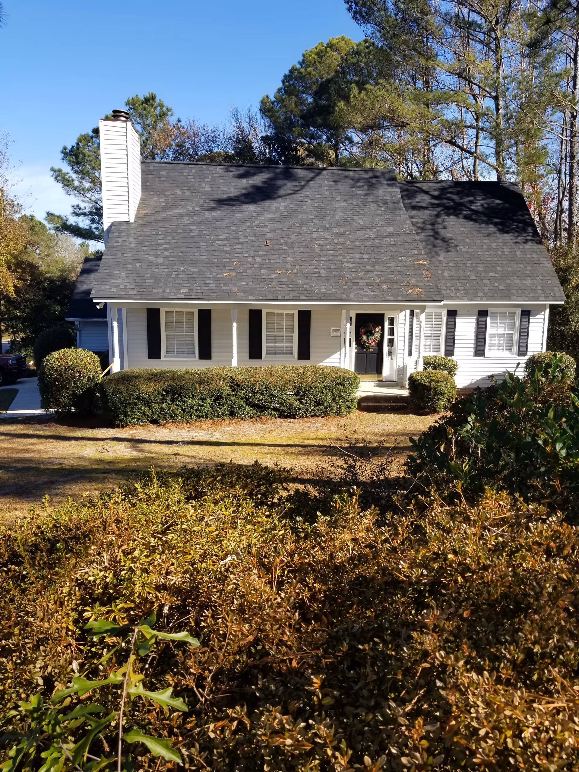 White single-story building with black shutters and a wreath on the front door, surrounded by trimmed bushes.