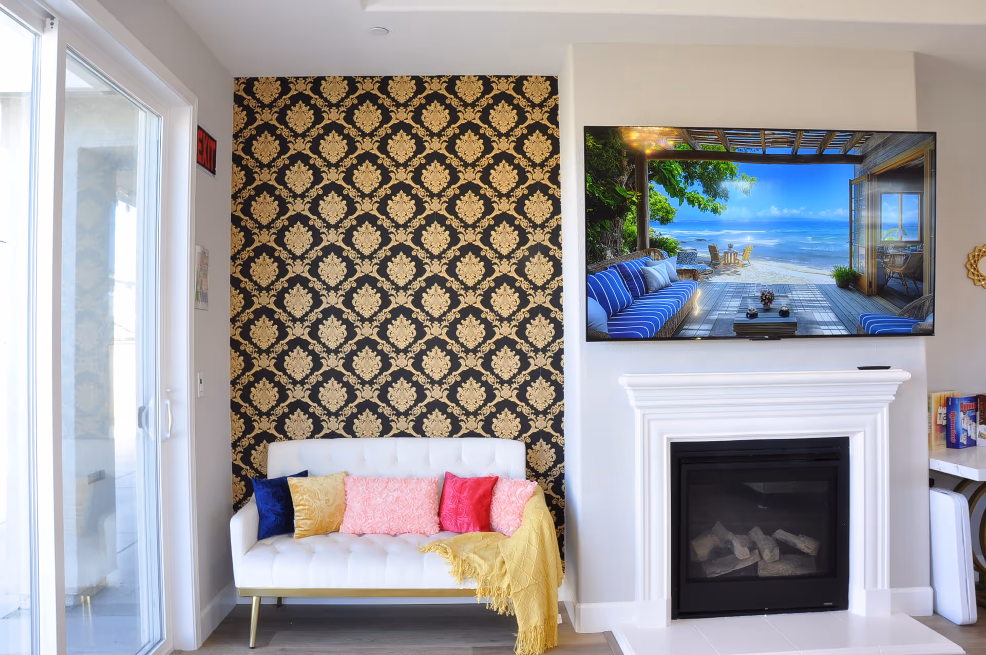 Bright living room with a white sofa and colorful pillows in front of a patterned accent wall, a wall-mounted TV above a white fireplace, and a sliding glass door.