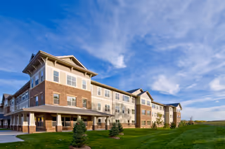 Exterior view of a large, multi-story senior living facility building with a combination of brick and light-colored siding under a partly cloudy blue sky. The building is surrounded by a well-maintained green lawn with small trees and shrubs.