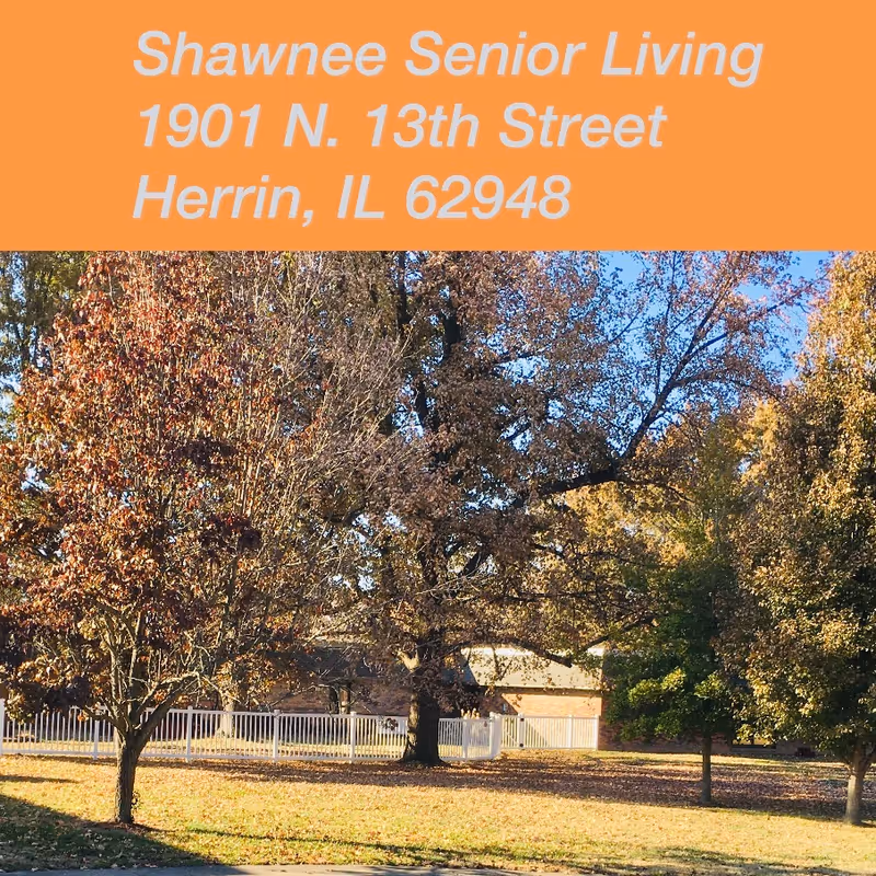 Outdoor view of Shawnee Senior Living facility showing trees with autumn foliage, a white fence, and a building partially visible in the background under a clear blue sky.