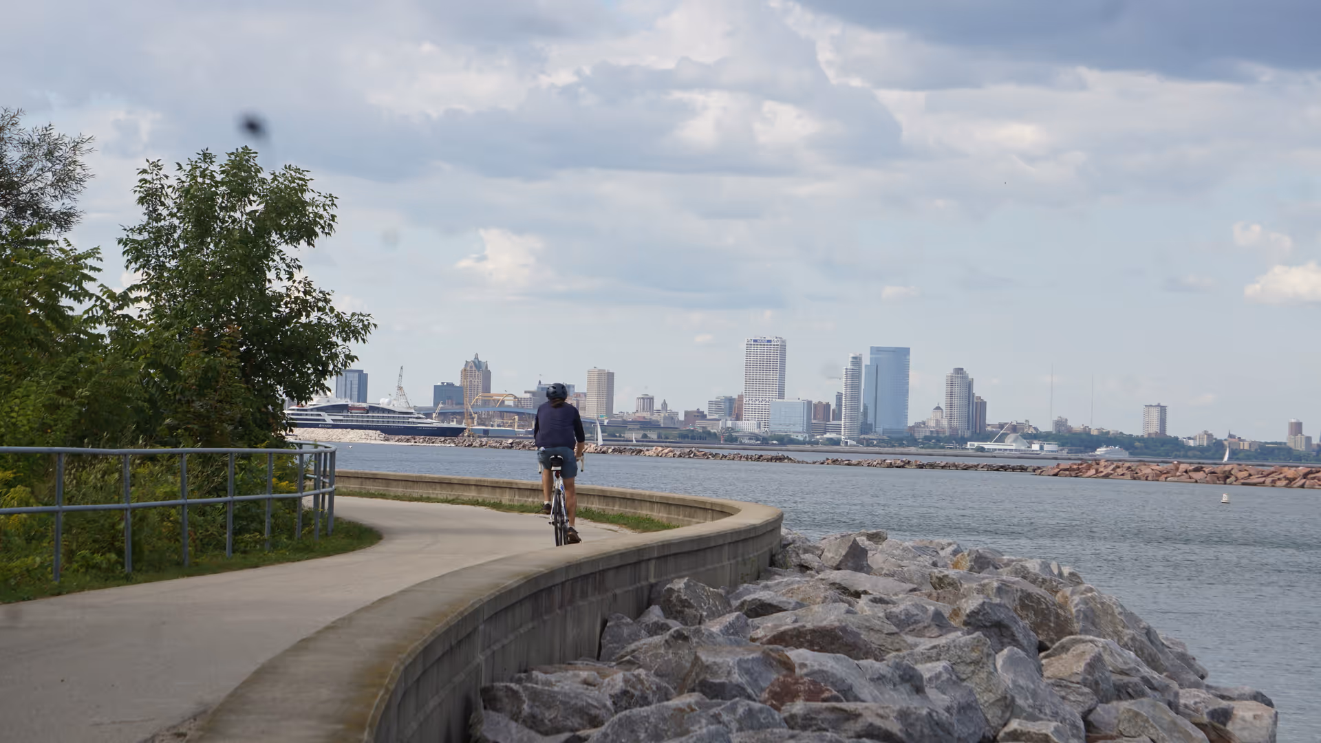 A cyclist rides along a curved waterfront path with a distant city skyline.