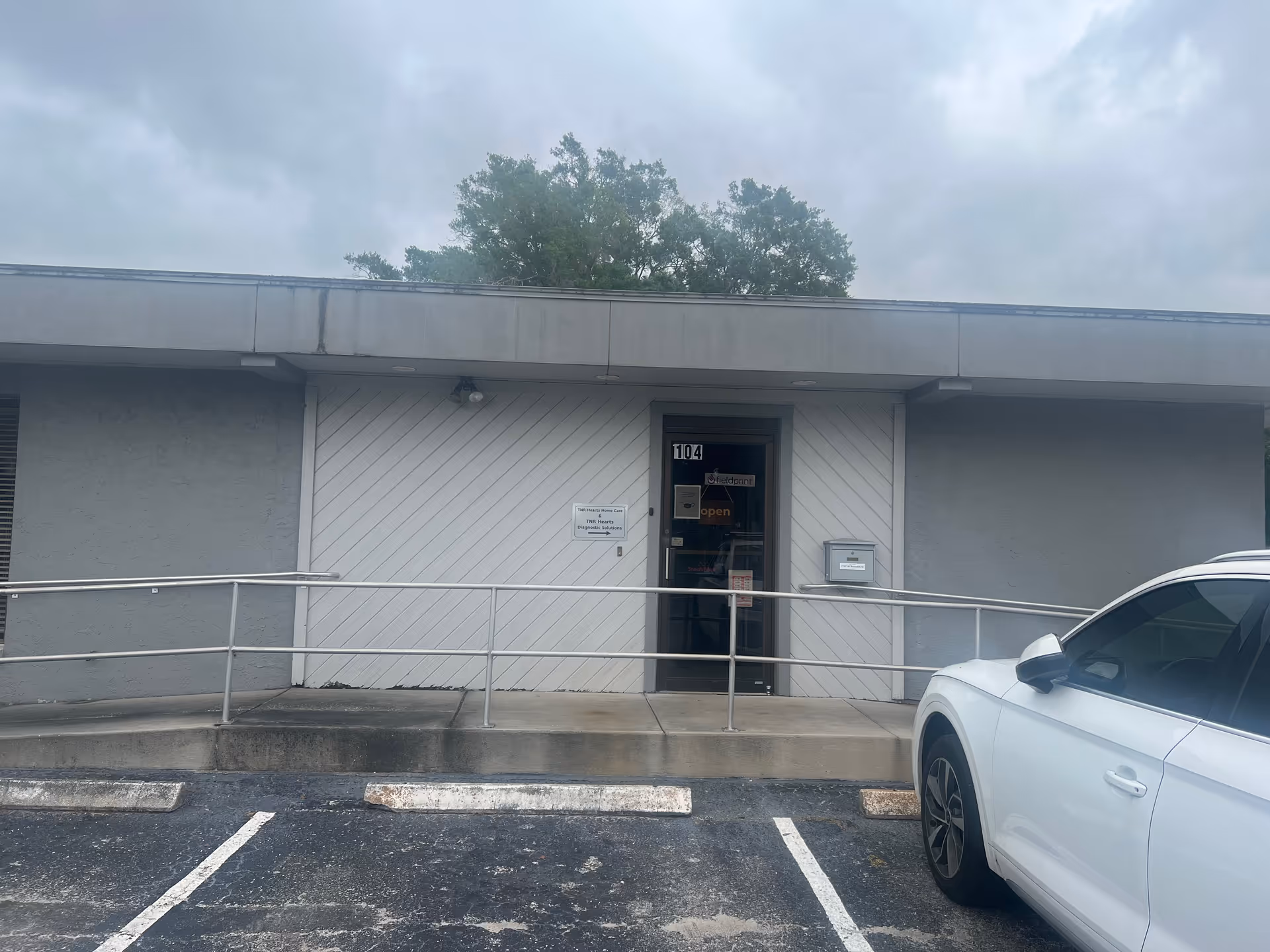 Front of a single-story facility with ramp, railing, glass door, and a parked white car.