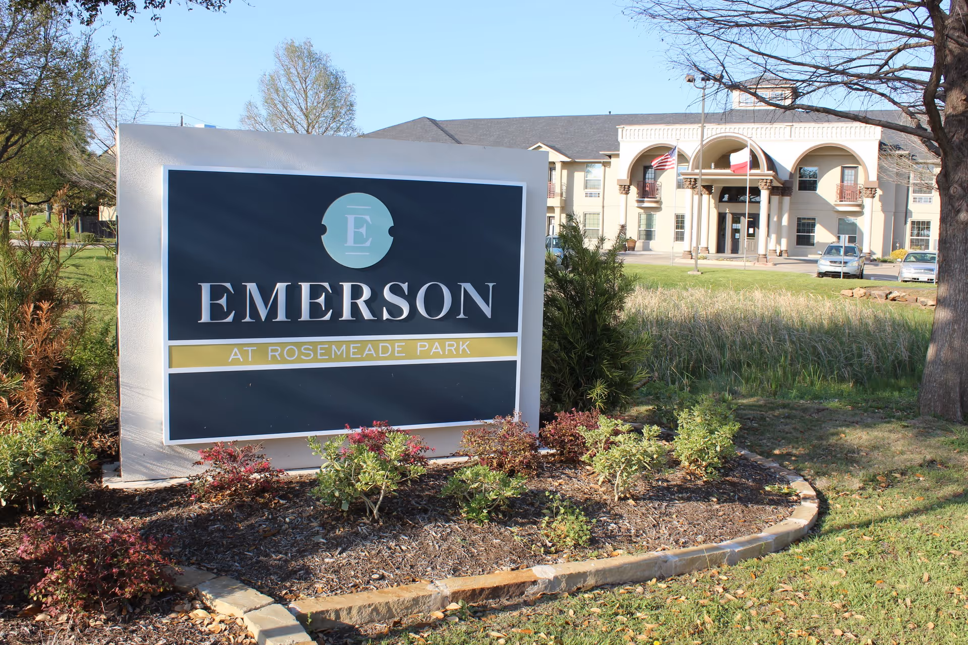 Outdoor view of Emerson at Rosemeade Park senior living facility sign with landscaping in front and the building with flags and parked cars in the background under a clear blue sky.