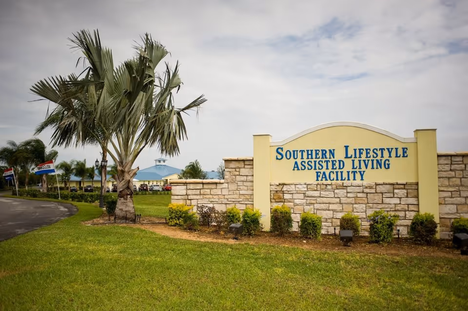 Exterior view of a senior living facility sign with stone wall and palm trees.